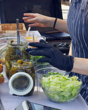 A gloved street food vendor reaches for fresh toppings among pickle jars and shredded lettuce at a Naplavka farmers market stall in Prague.