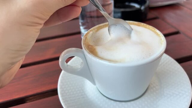 Woman's hand slowly stirring cappuccino foam with a teaspoon in an elegant white cup