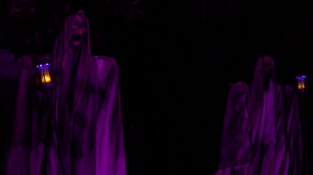 Halloween yard scene shows two hooded ghosts holding lanterns in purple light at night. Spooky outdoor decorations with draped fabric, skull faces, and dark trees.
