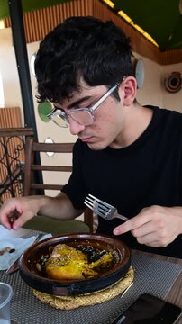 Marrakech, morocco. August 8, 2026. Young man with glasses eating traditional moroccan chicken tagine with prunes and sesame seeds. Culinary travel experience in a marrakech restaurant