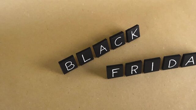 Black tiles with handwritten white letters forming the words Black Friday gliding from right to left over a light brown paperboard background, ideal for announcing sales and promotions