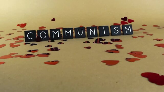 Gliding shot showing the word communism written with black tiles on a light brown background, surrounded by scattered red confetti in the shape of hearts, symbolizing political ideology