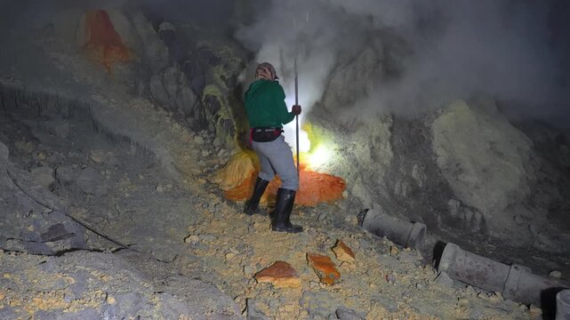 Sulfur miner chisels a large sulfur block amid dense volcanic fumes inside Kawah Ijen in a static handheld shot.