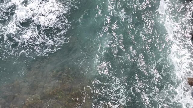 Top-down aerial view of Atlantic seawater near Asilah, Morocco, showing swirling foam, shallow submerged rocks and textured wave patterns along the coastal waters.