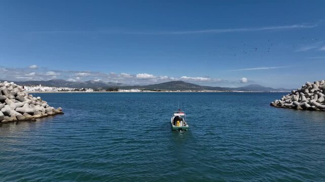 Aerial view of a small boat navigating through a protected Mediterranean harbor entrance in Northern Morocco, passing between concrete breakwaters with distant mountains and coastal scenery in the bac
