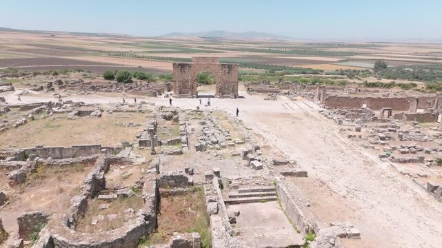 Wide aerial view across the archaeological site of Volubilis in Morocco, showing extensive ancient ruins, foundations and streets leading toward the Arch of Caracalla, surrounded by open agricultural