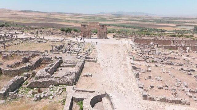 Wide aerial view along the ancient Roman street at Volubilis, Morocco, leading toward the Arch of Caracalla and surrounded by excavated building foundations and archaeological ruins.