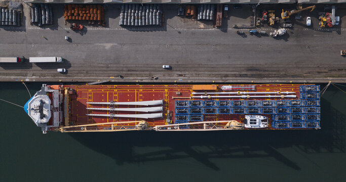 Portugal - 22 August 2026: Aerial view of a cargo ship docked at a harbour, loaded with large white wind turbine blades and industrial equipment, with trucks parked on the quay.
