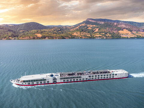 Aerial diagonal view of a river cruise ship cutting a white wake through broad blue waters toward rolling hills at sunset - Danube, Iron Gates Region.