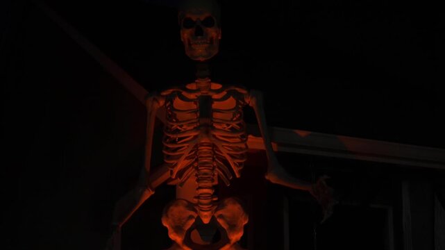 Large skeleton decoration glows orange against a dark suburban house. Bones, ribs, and skull are highlighted with dramatic light for a spooky Halloween scene.