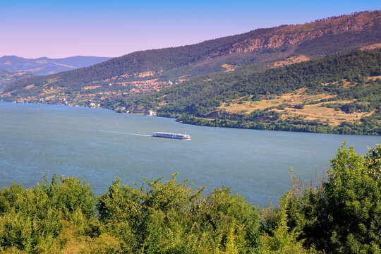 Bright summer light over the wide Danube River as a cruise ship glides through a green mountain gorge.