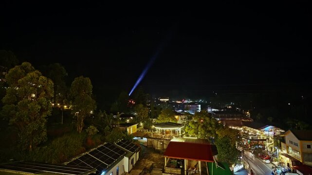 A narrow blue searchlight sweeps the dark sky over Ella as warm street lamps and shopfronts illuminate the small hill country town below in Sri Lanka.