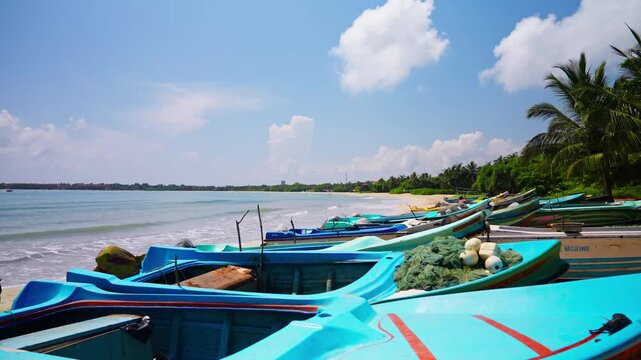 Brightly painted wooden fishing boats rest in a long row on golden sand beside the turquoise sea, with coconut palms and a distant shoreline on the coast of Sri Lanka, the fleet of a fishing village.