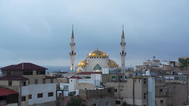 King Hussein bin Talal Mosque above central Madaba, Jordan, with a golden dome and twin minarets rising over dense urban rooftops beneath blue evening skies, timelapse