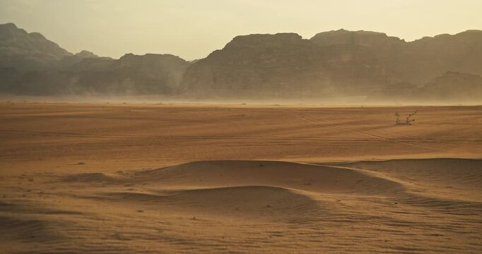 Golden sand dunes stretch across the vast Wadi Rum desert in Jordan, with windblown dust, rippled sand and rugged rocky mountains rising in the distance.