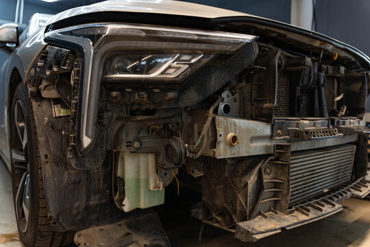 Damaged vehicle front end with exposed headlight, grille area, and missing bumper parts. Dusty bodywork reveals internal brackets, wiring