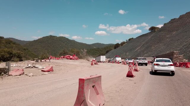 Road construction zone in Huatulco Oaxaca showing vehicles driving beside traffic barriers and a rocky hillside under a clear blue sky during roadwork in Mexico