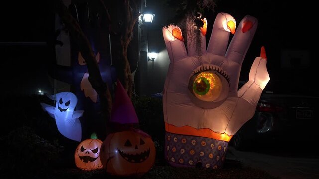 Close-up Halloween yard display at night with a giant inflatable hand with an eyeball, glowing ghosts, and jack-o'-lanterns. A suburban house and driveway show festive lights and decorations for trick