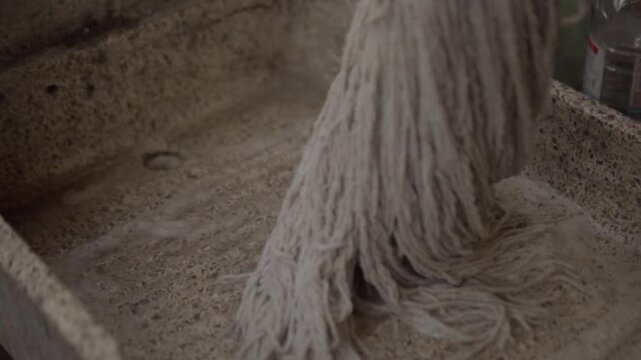 Close up view of mop being squeezed against textured stone floor in outdoor patio. White strands compress together as the mop head is pressed firmly against the rough surface.