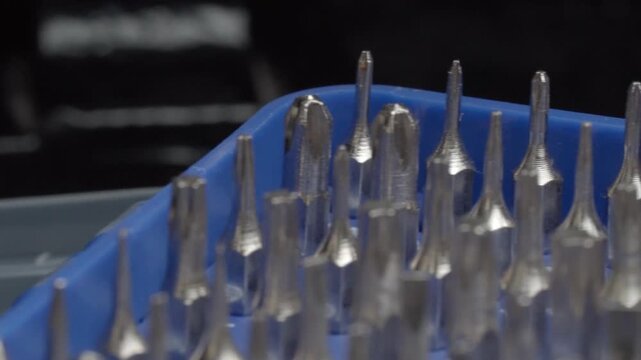 Close up view of metal drill bits arranged in blue plastic container on workbench. Various sizes and shapes of shiny steel bits are displayed together in detailed macro view