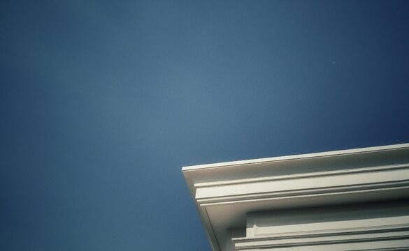 Minimalist white architecture cornice corner against a clear blue sky. Artistic low angle shot with vintage analog film style photography, subtle grain texture, and nostalgic mood.