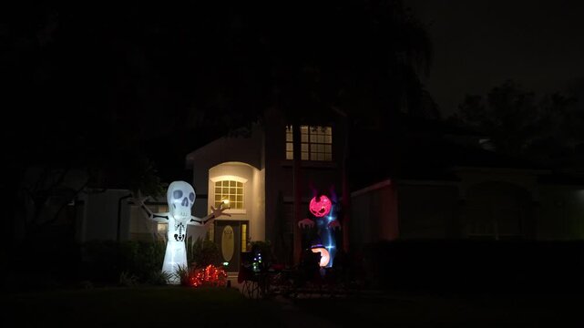 Suburban house at night with a tall inflatable skeleton and a glowing demon pumpkin figure in the yard. Porch lights, windows, and lawn decorations create a spooky neighborhood scene.