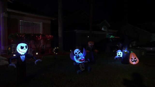 Suburban house at night with a Halloween yard full of glowing inflatables, pumpkins, and a spooky tree. Ghosts and skeleton lights decorate the lawn by parked cars.