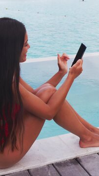 Phone lifestyle. Woman using mobile phone relaxing in the sun by pool on beach in on vacation. Lady using phone app at luxury travel holidays resort. Raw vertical video of real woman without makeup