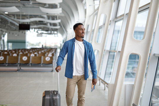 A young Black man with a suitcase and passport walks through a bright, contemporary airport terminal with rows of seats and large windows