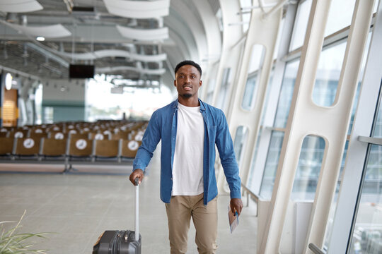 A smiling man in casual attire pulls his luggage through a bright, contemporary airport concourse with large windows and seating areas