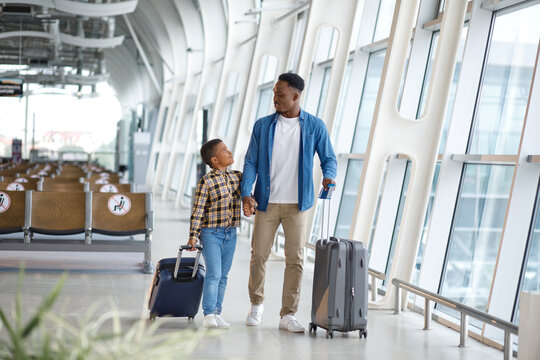 A father and his young son walk together through a modern airport terminal, pulling their suitcases and looking at each other