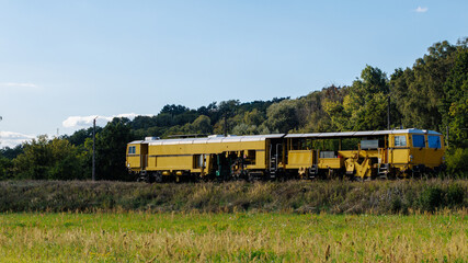 A long yellow railway maintenance train parked on tracks against a dense green forest under a clear blue summer sky. © Dmytro
