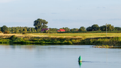 Obraz premium A green navigation buoy floats in a calm river with village houses and green meadows under a blue sky in summer.