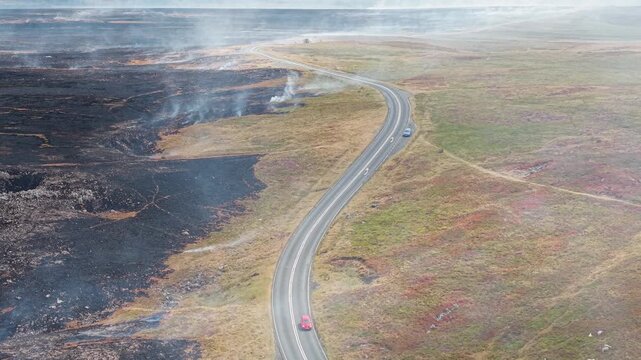 Red car moving along country road through burnt moorland, smoke drifting over burnt heather and grass after hill fire in Brecon Beacons National Park