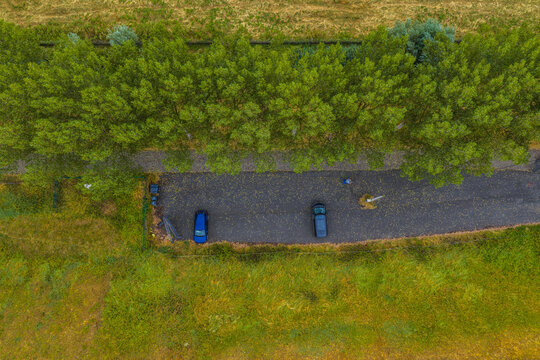 Beveren-Kruibeke-Zwijndrecht, Belgium - 18 August 2026: Aerial view of a small parking area with two cars parked on asphalt, bordered by a dense row of green trees and surrounding grassy fields.