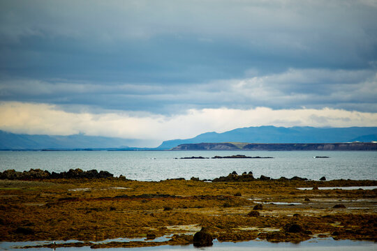 Seaweed covered basalt reefs at low tide in coastal bay with view of distant Melarsveit coastline and hills under moody cloudy sky in Akranes Iceland