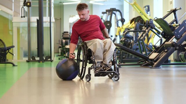 Positive confident disabled man in wheelchair working out with heavy medical ball at training in gym, strong injured sportsman lifting and throwing slam ball, workout for upper body muscle strength.