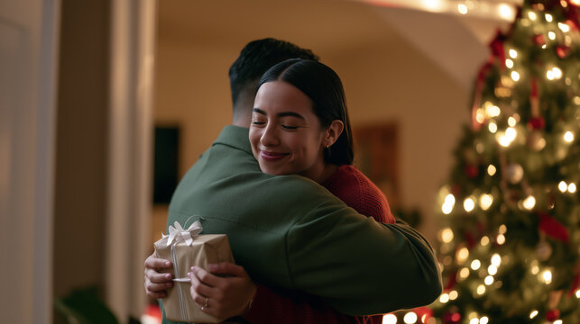 Christmas couple embracing with affection and gratitude while holding a wrapped gift beside a decorated Christmas tree. 
