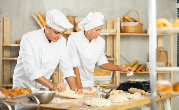 Two young bakers prepare dough in kitchen of bakery - together they knead dough, cut it with a knife and weigh it on scales