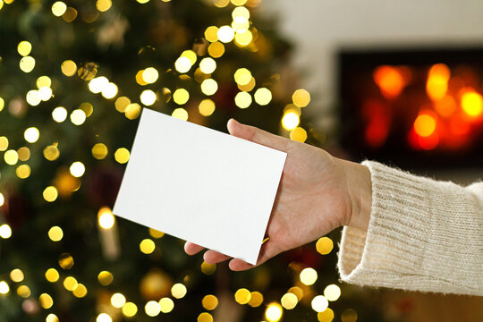 Woman holding blank white greeting card mockup in hands on cozy bokeh lights background, concept of Christmas invitation, wishlist, holiday greetings and copy space
