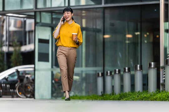 Young professional woman walking with coffee and talking on phone outside modern office building