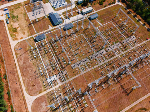 Top down aerial view of power substation. Metallic frames and transmission components cast long shadows on brown soil and paved access paths.