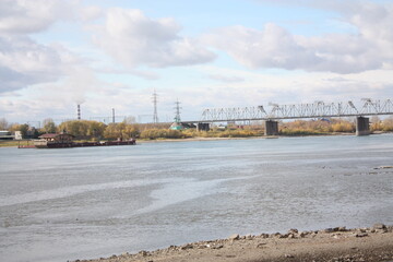 Naklejka premium A wide landscape of a broad river with a rocky shoreline on the foreground, featuring a metal bridge and transmission towers in the distance.