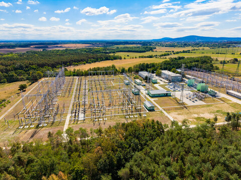 Overhead view of an electrical power grid distribution station. Metal switchgear structures and transformers rest on green ground surrounded by pine trees under a blue sky.