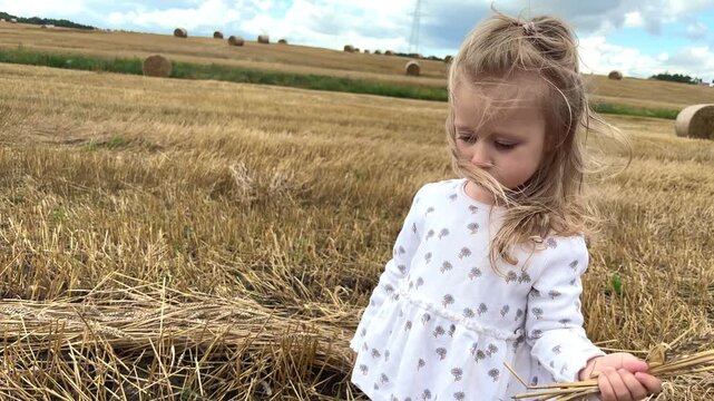 Young girl shows a small bouquet of wheat and straw stalks in a harvested field, rural childhood, agriculture, summer harvest, countryside lifestyle.