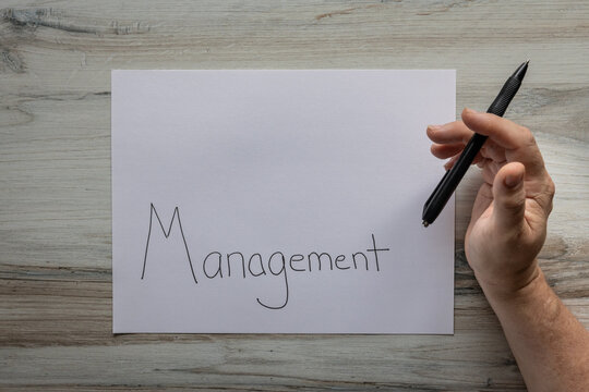 Flat lay background white sheet of paper with negative space available with the word Management handwritten on it with a hand holding a black pen.