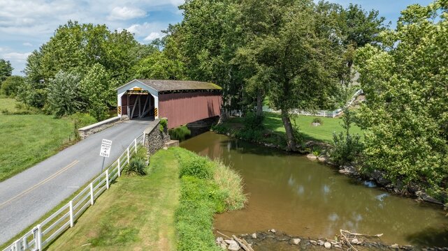 Aerial view of a historic red covered bridge spanning a calm stream surrounded by lush green trees and a white fence in Lititz, Pennsylvania, United States.