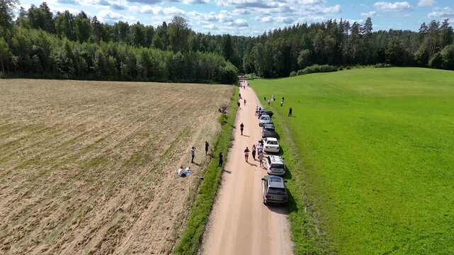 Distance runners move along a gravel farm road lined with parked cars and spectators on a sunny rural marathon course.