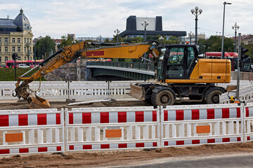 Yellow wheeled excavator on urban road construction site surrounded by safety barriers near bridge in city center. Urban renewal, road repair, public works © A-Quantix