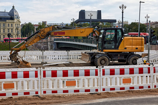 Yellow wheeled excavator on urban road construction site surrounded by safety barriers near bridge in city center. Urban renewal, road repair, public works